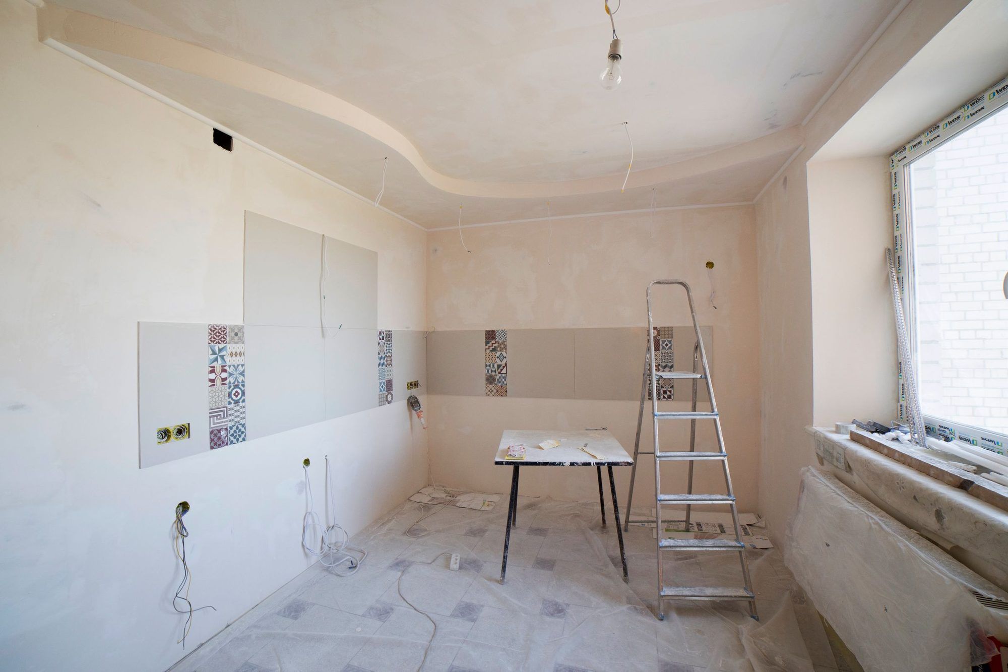 Cocina de una vivienda a medio reformar, con el alicatado a medias, la instalación eléctrica vista, una escalera y el suelo protegido