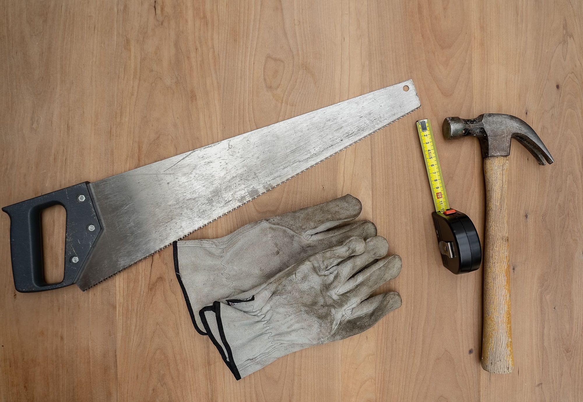 Serrucho, guantes de trabajo, flexómetro y martillo sobre una mesa de madera